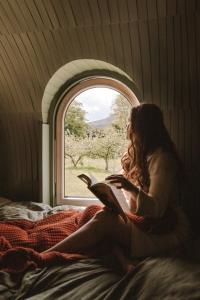 a woman sitting on a bed reading a book at Enniskeen Estate & Forest Spa - Boutique Riverside Glamping Cabins in Newcastle