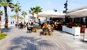 people sitting at tables and chairs on a sidewalk with palm trees at beach front tel aviv bat yam apartment -hotel 39 in Bat Yam