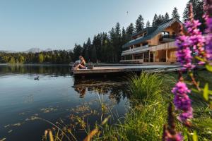 une femme assise sur un quai à côté d'une maison sur un lac dans l'établissement Alpenhotel Kitzbühel am Schwarzsee - 4 Sterne Superior, à Kitzbühel