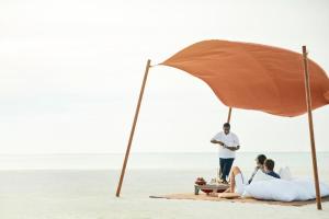 three people sitting under an umbrella on the beach at COMO Cocoa Island - Partner Travels FREE for 7 Nights or More in Male City