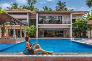a woman sitting next to a swimming pool in front of a house at Saffronstays Casa Del Palms, Alibaug - luxury pool villa with chic interiors, alfresco dining and island bar in Alibaug