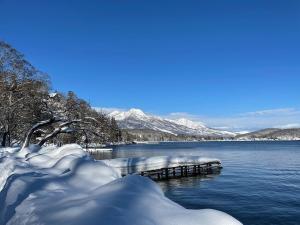 a snowy lake with a dock and snow covered mountains at Myoko Woodland Lodge in Myoko