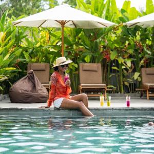 a woman in a hat sitting next to a swimming pool at Bruga Villas Restaurant and Spa in Selong Belanak