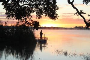 een man die vist op een dok op een meer bij zonsondergang bij Kayube Zambezi River House in Livingstone