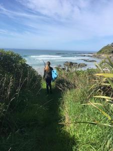a woman walking down a path near the beach with a surfboard at Stay in School by the Sea in Dunedin