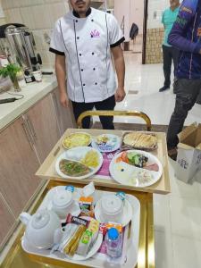 a chef standing in a kitchen with two trays of food at Al Farhan Apartment (Al Jubail-Al Balad ) in Al Jubail