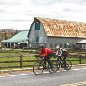 two men riding bikes down a road near a barn at Domaine Dorchamps in Lac-Brome