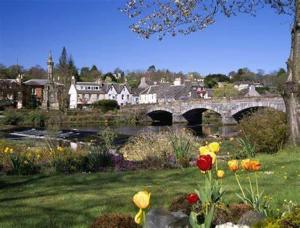 eine Brücke über einen Fluss mit Blumen im Hof in der Unterkunft HV66 - 3 Bedrooms Indoor Pool Loch Views Fishing Golf Riding Shooting Water Sports 15 Mins Drive To Beaches PASSES NOT INCLUDED Most Activities Will Not Be Available Out Of Season Please Check Before Booking in Newton Stewart
