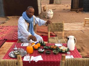 un hombre sentado en una mesa con comida. en Tikida Camp by tinfou en Zagora