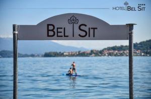 two people on a surfboard in the water under a sign at Hotel Bel Sit in Meina