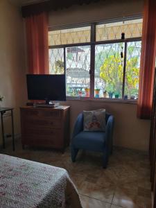 um quarto com uma televisão, uma cadeira e janelas em Casa aconchegante em Vila Valqueire no Rio de Janeiro