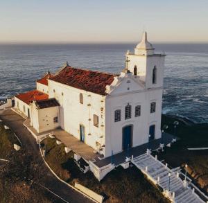 Una vieja iglesia blanca con el océano detrás. en Nosso Repouso Saquarema - Casa inteira com Piscina,churrasqueira privativos, Wi-fi,900m da praia, Tv-Smart. en Saquarema