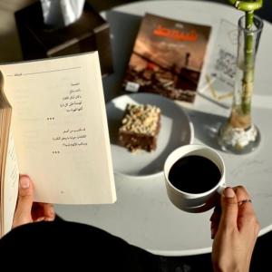 une personne assise à une table avec une tasse de café et un livre dans l'établissement The District Hotel Najran, à Najran