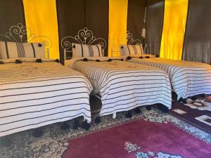 three beds in a room with yellow curtains and a rug at Bivouac Draa in Zagora