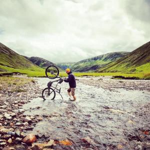 a man carrying a bike across a river at Baltinna East - by Where Stags Roar in Newtonmore