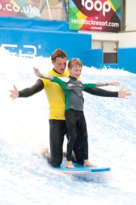 two boys are standing on a surfboard in the water at Harlyn lodge Hot Tub Lodge at Retallack Resort in Saint Columb Major