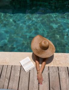 une femme avec un chapeau et un livre à côté d'une piscine dans l'établissement La Siesta bungalows & Cuisine, à Gili Air