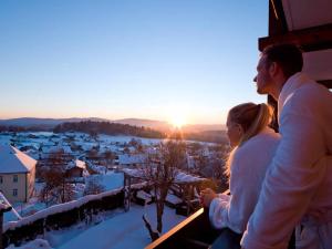 un hombre y una mujer de pie en un balcón viendo la puesta de sol en Panoramahotel Grobauer, en Spiegelau