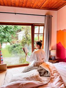 a woman sitting in bed reading a book in front of a window at Capivari Lodge vista incrível da natureza in Campos do Jordão