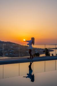 Le Ciel Suites & Villas في مدينة ميكونوس: فتاة تقف على حافة مع غروب الشمس في الخلفية
