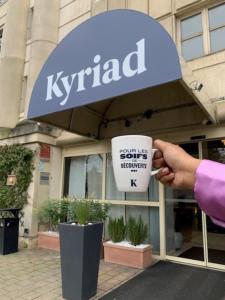 una persona sosteniendo una taza de café frente a una tienda de kirrip en Kyriad Hotel Montpellier Centre Antigone, en Montpellier