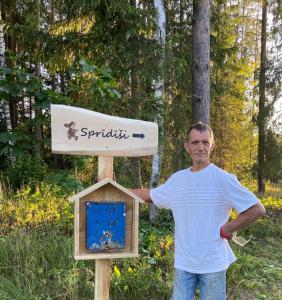 Un uomo in piedi accanto a un cartello con una casetta per uccelli di Sprīdīši 