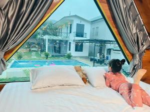 Una niña acostada en una cama mirando por la ventana hacia una piscina. en Cloudy Home - Venuestay, en Hanói