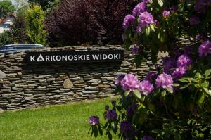 a sign on a stone wall with purple flowers at Karkonoskie Widoki - Jacuzzi z widokiem na Śnieżkę - pyszne śniadania i parking w cenie - pokoje z balkonami in Karpacz