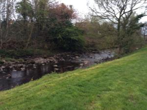 a grassy hill next to a river with trees and a field at Corner House 51 Go Go Street Ground Floor Largs KA308JW 2 Bedroom in Largs