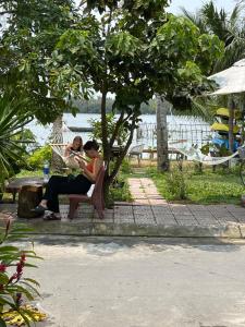 two people sitting on a bench under a tree at Coral Riverside Homestay & Hostel in Hoi An