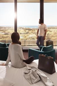 a woman sitting on a bed looking out the window at a man at RACV Torquay Resort in Torquay