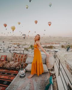 una mujer con un vestido naranja parada en un balcón con globos de aire caliente en Seki Cave Suites, en Göreme