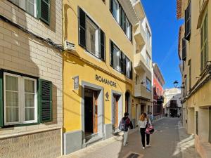 twee mensen die door een straat lopen voor een geel gebouw bij ROMANDRE in Alaior