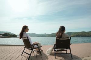 dos mujeres sentadas en sillas por un cuerpo de agua en TADAYOI 海士グランピング, 