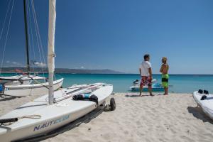 two people standing on a beach with boats in the water at Hotel Dei Pini in Alghero