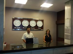 un homme et une femme debout derrière un comptoir avec horloges dans l'établissement Fenicia Palace Hotel, à Varginha