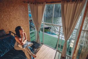 uma mulher sentada numa cama num quarto com uma janela em แพคิงส์ราฟ เขื่อนศรีนครินทร์ กาญจนบุรี em Ban Wang Khun Knachen
