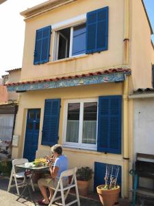 a person sitting at a table outside of a house with blue shutters at Cabanon aux Goudes Marseille calanques in Marseille