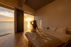 a girl sitting on a bed looking out a window at the ocean at 湘南OCEAN HOUSE in Koshigoe