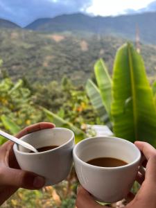 two hands holding cups of coffee in front of a mountain at Refugio de Mery Lucmabamba in Sahuayacu