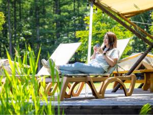 a woman sitting in a chair on a deck with her cell phone at Tokachi Nakasatsunai Glamping Resort Feriendorf in Naka-satsunai