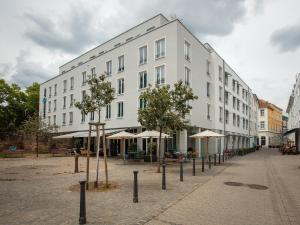 un bâtiment blanc avec des arbres et des parasols dans une rue dans l'établissement Motel One Saarbrücken, à Sarrebruck