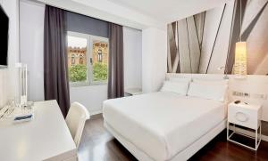 a white bedroom with a bed and a desk and a window at NH Barcelona Stadium in Barcelona