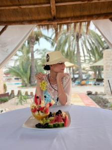 a woman wearing a hat sitting at a table with a plate of fruit at Le Patio de Mezraya in Mezraya