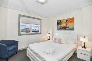 a white bedroom with a bed and a blue chair at Sandy Cove Apartment The Entrance 24 in The Entrance