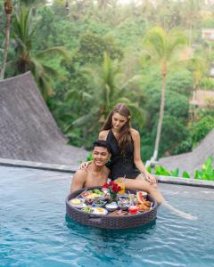 Ein Mann und eine Frau sitzen auf einem Tablett mit Essen im Pool. in der Unterkunft LeRosa Valley Resort in Ubud