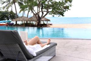 a woman laying on a chair reading a book next to a pool at Deva Beach Resort Samui in Choeng Mon Beach