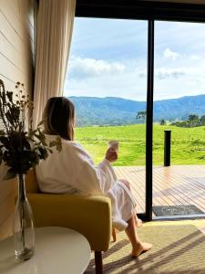 a woman sitting on a couch with a cup of coffee looking out the window at Refúgio Serra e Flor in Urubici