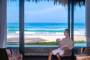 a woman sitting in a chair holding a coffee cup in front of the beach at Sealord Naithon Beachfront Villa in Nai Thon Beach