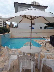 uma mesa branca e cadeiras com um guarda-sol ao lado de uma piscina em Apto Enseada Coral Gables no Guarujá